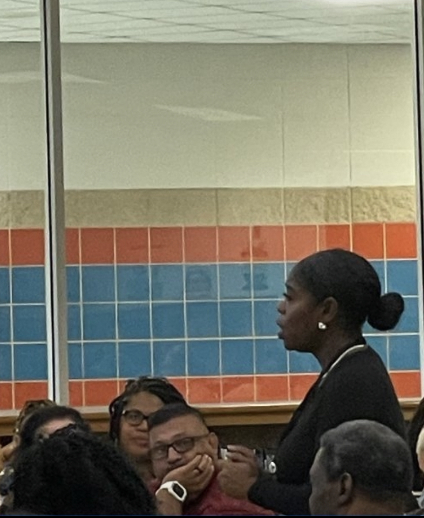 Woman speaking to a group of people in a room with tiled walls, some people wearing glasses, and a window behind her.