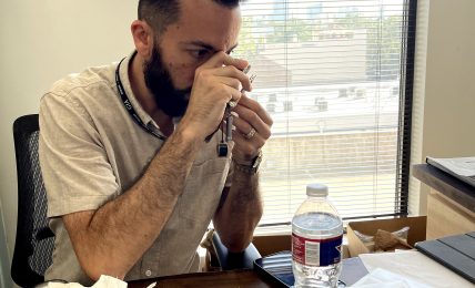 Man sitting at desk, drinking from a cup, with a window behind him showing a cityscape, office supplies on desk.