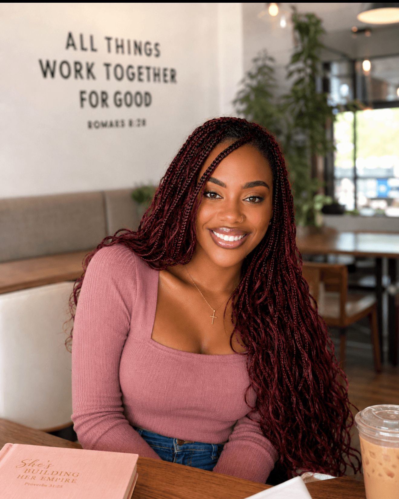 Smiling woman with long braided hair sitting in a cafe, wearing a pink top, with a quote on the wall behind her.
