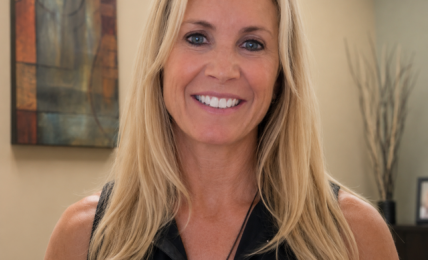 Smiling woman with long blonde hair in a black sleeveless top, standing indoors with artwork and plants in background.
