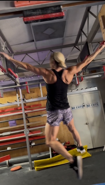 Person with blonde hair in a black tank top and patterned shorts balancing on a yellow board with arms outstretched.
