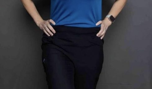 Woman standing with hands on hips, smiling, wearing a blue shirt and black pants, against a gray background.