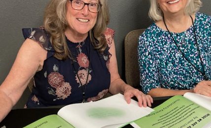 Two women sitting at a table with open books and papers, smiling, in a gray room.