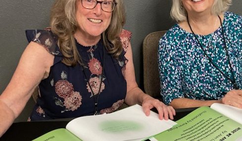 Two women sitting at a table with open books and papers, smiling, in a gray room.