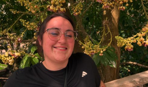 Smiling woman with glasses and black athletic shirt standing outdoors near a wooden railing, surrounded by yellow flowering tree branches.