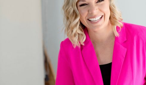 Smiling woman with blonde wavy hair wearing a bright pink blazer, standing indoors against a light background.