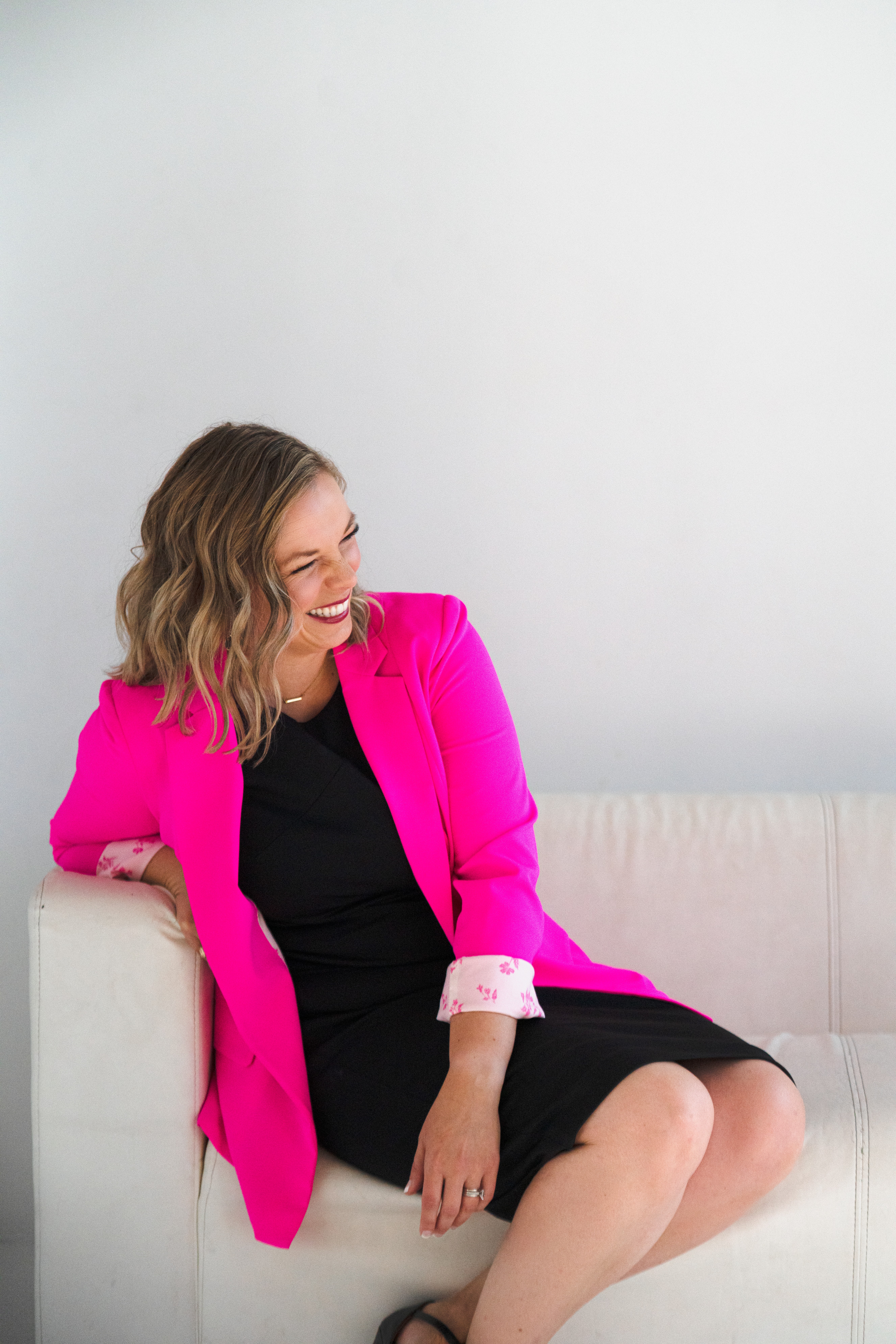 Woman with wavy hair wearing a pink blazer and black dress, sitting on a white sofa, smiling and looking to the side.