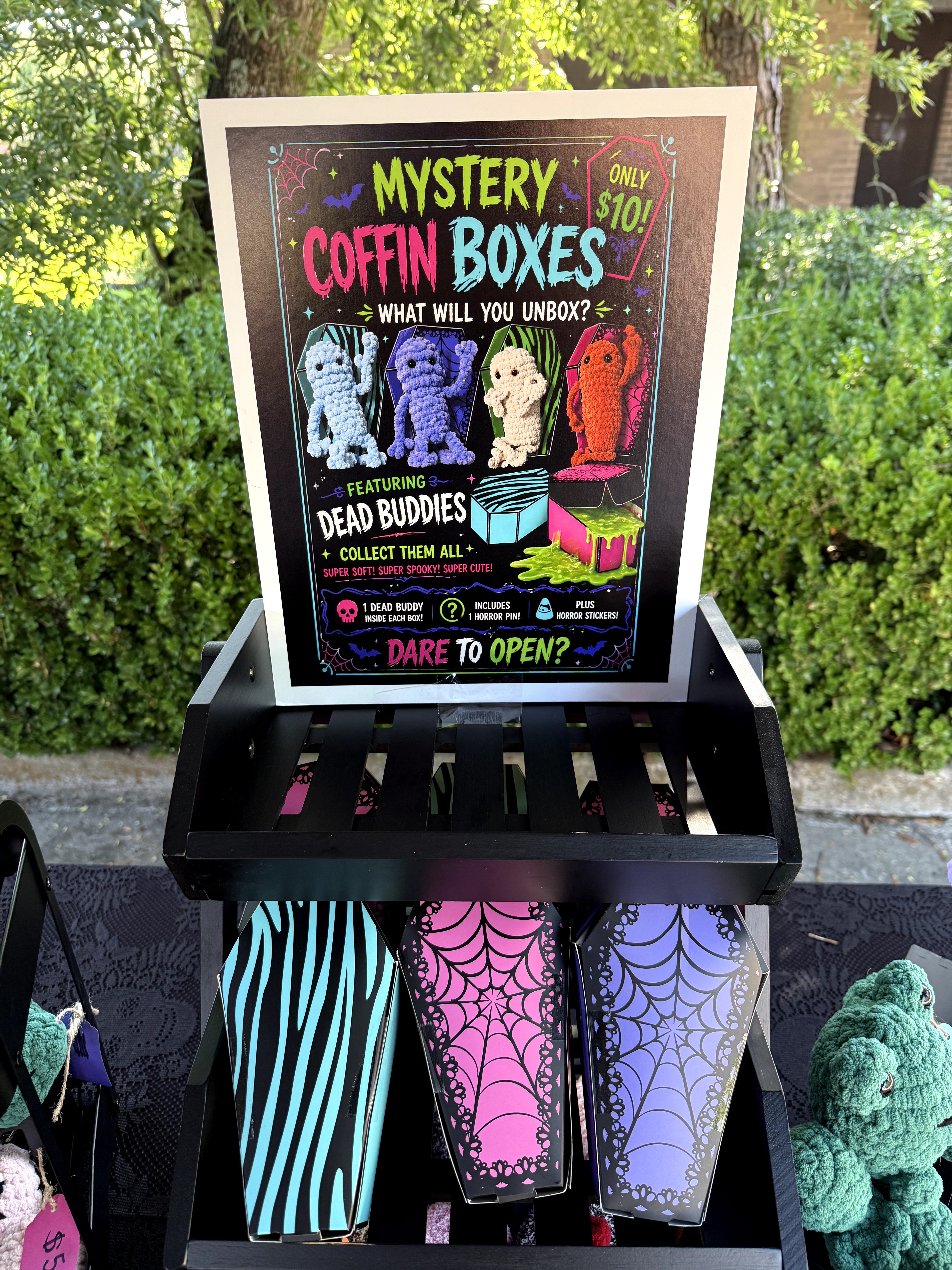 A colorful chalkboard sign advertising mystery coffin boxes with cartoon characters, set outdoors with greenery in the background.