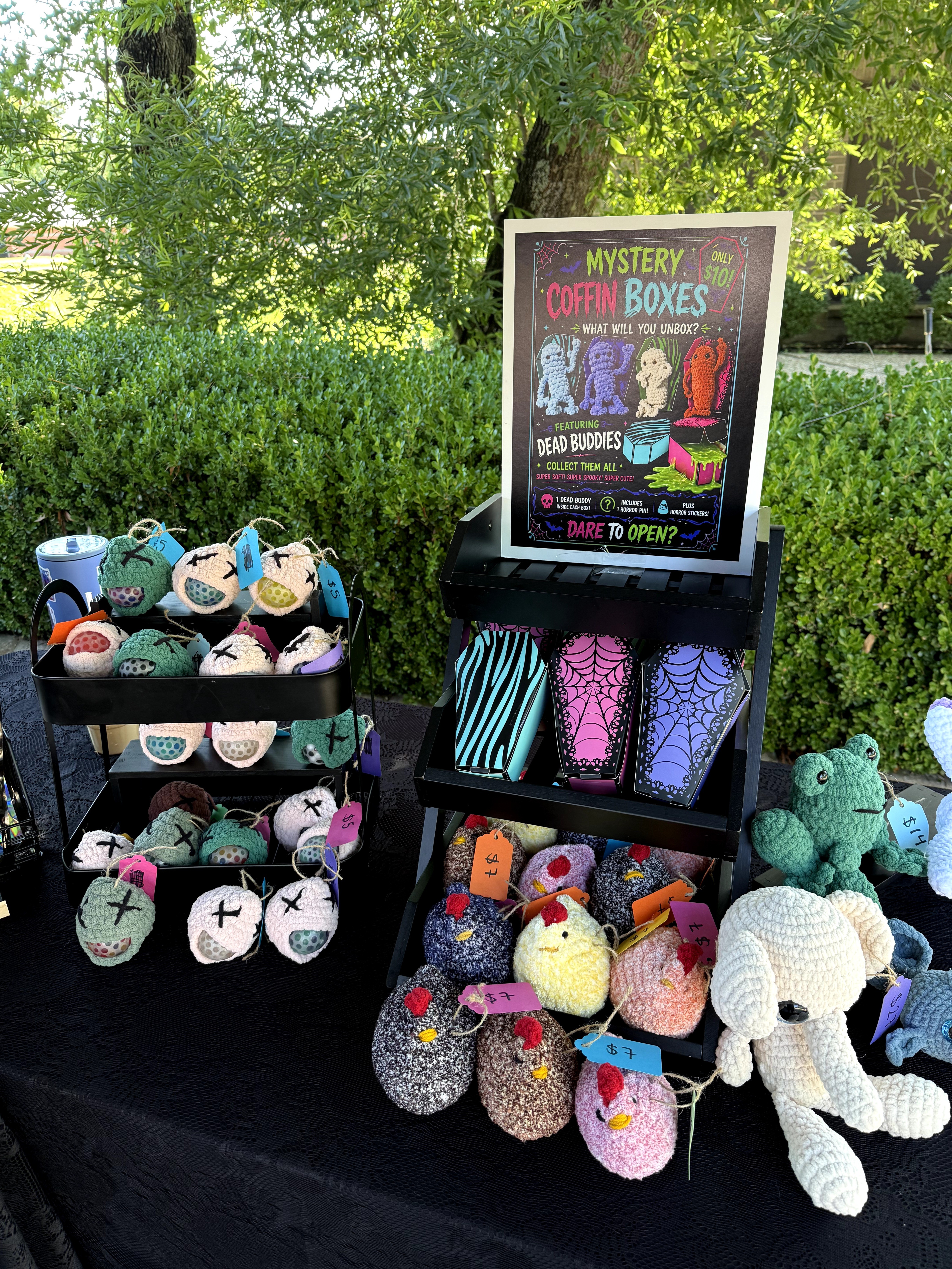 Display of colorful crocheted stuffed animals and boxes outdoors with a sign about mystery cotton boxes, surrounded by greenery.