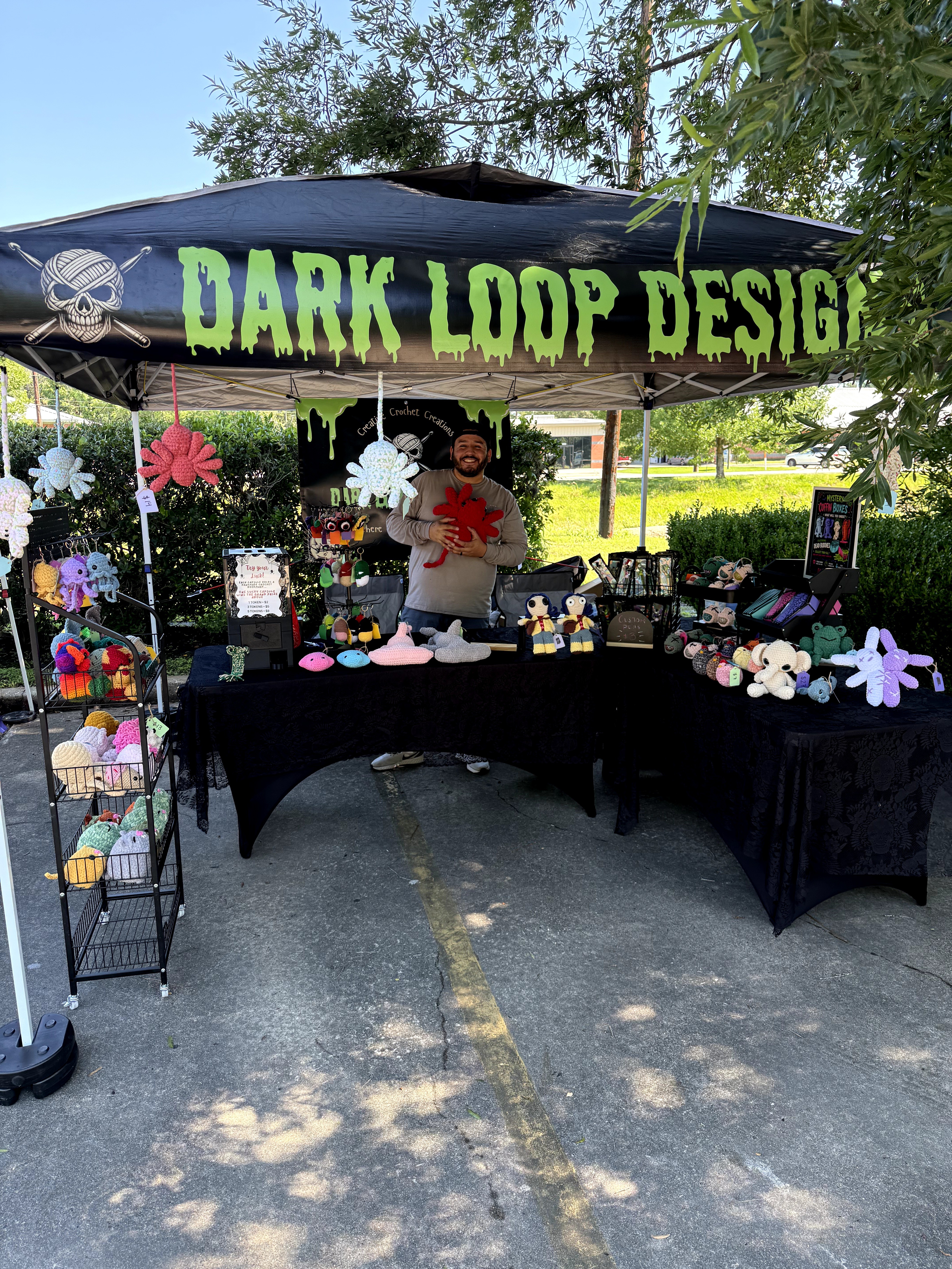 Person standing behind a table with colorful items under a black canopy with green text, outdoors with trees and sunlight.