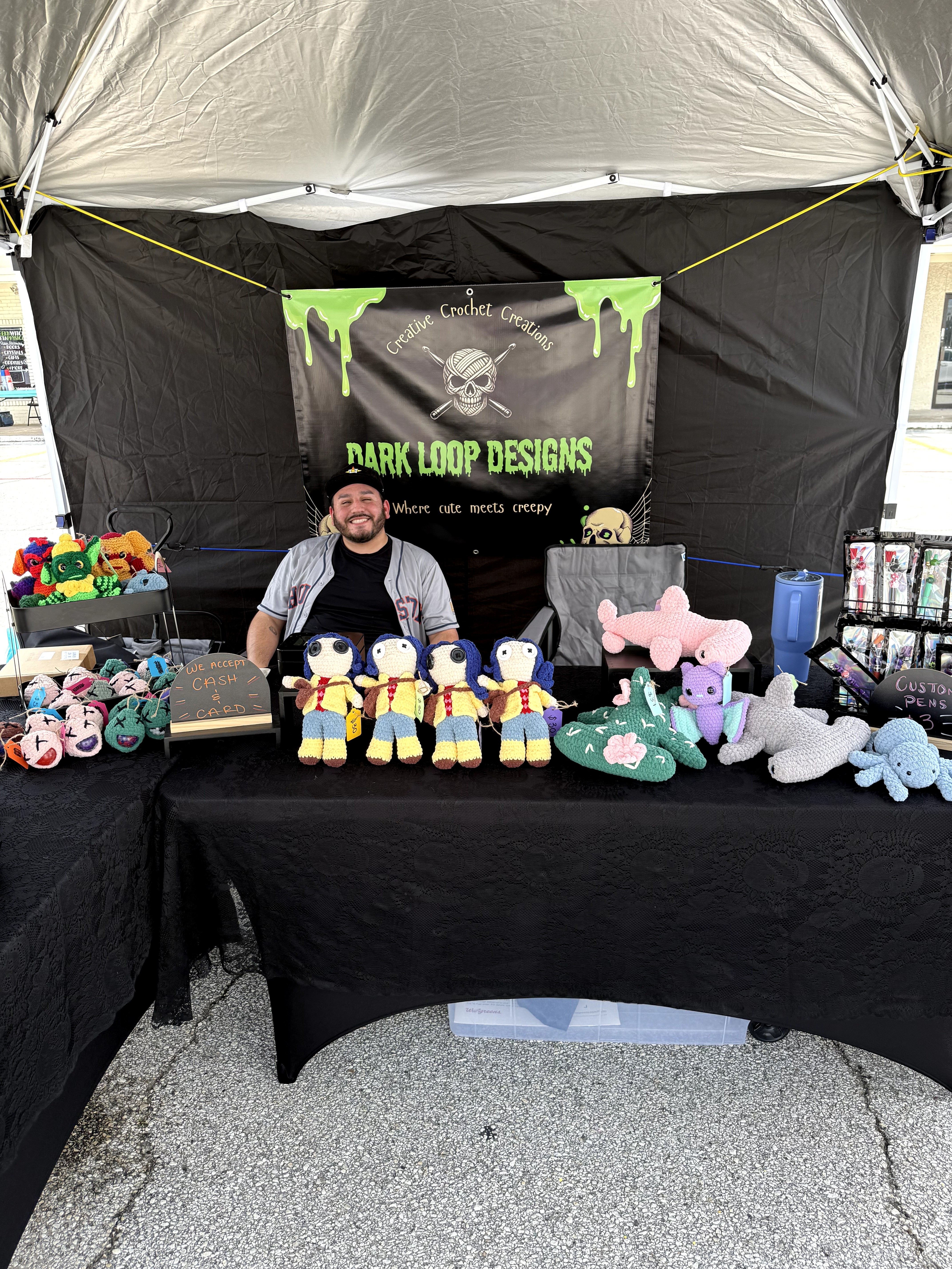 Person sitting at a booth with plush toys and a banner reading 'Dark Loop Designs' under a tent.