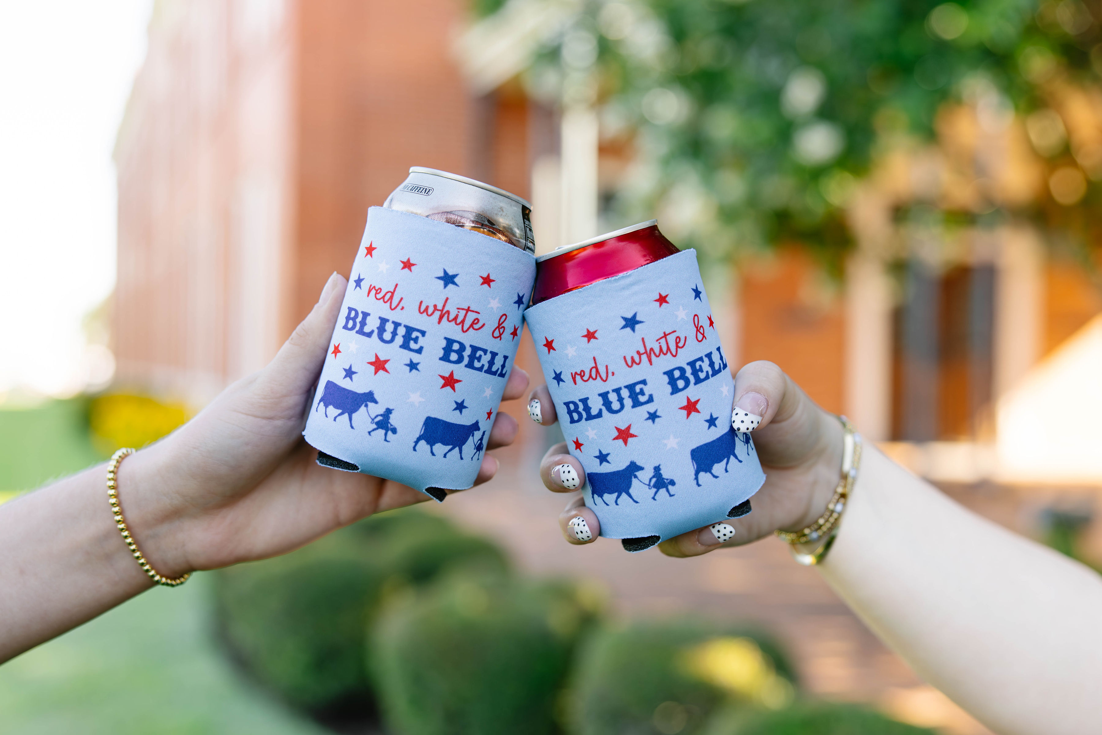 Two hands holding cans with patriotic blue, red, and white designs, outdoors with blurred greenery and building background.