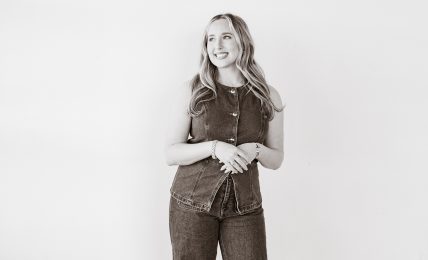 Young woman with long hair standing against a plain white wall, smiling, wearing a sleeveless top and wide-leg jeans, hands clasped.