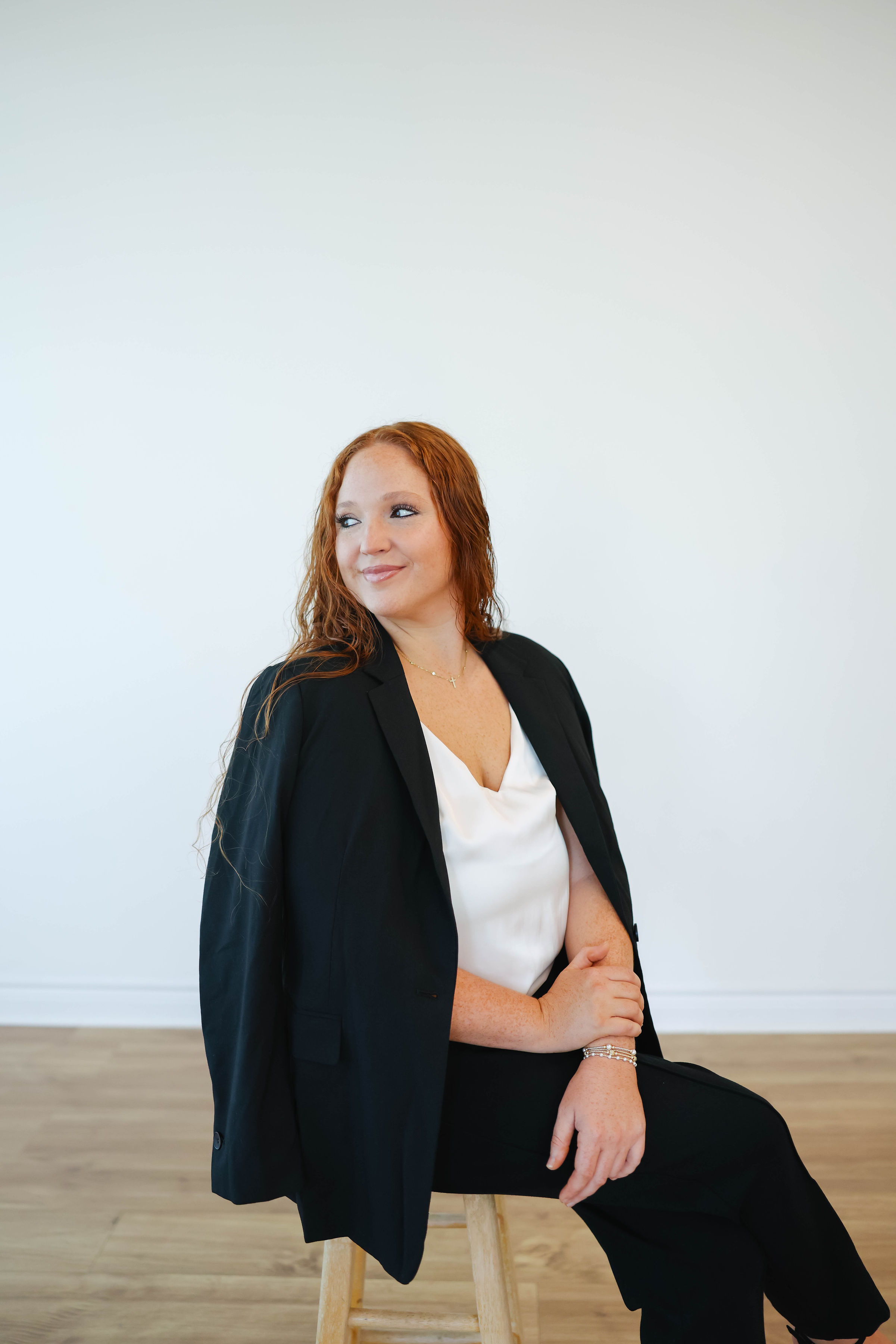 Woman with red hair sitting on a stool, wearing a black blazer and white top, looking to the side, against a plain white wall.