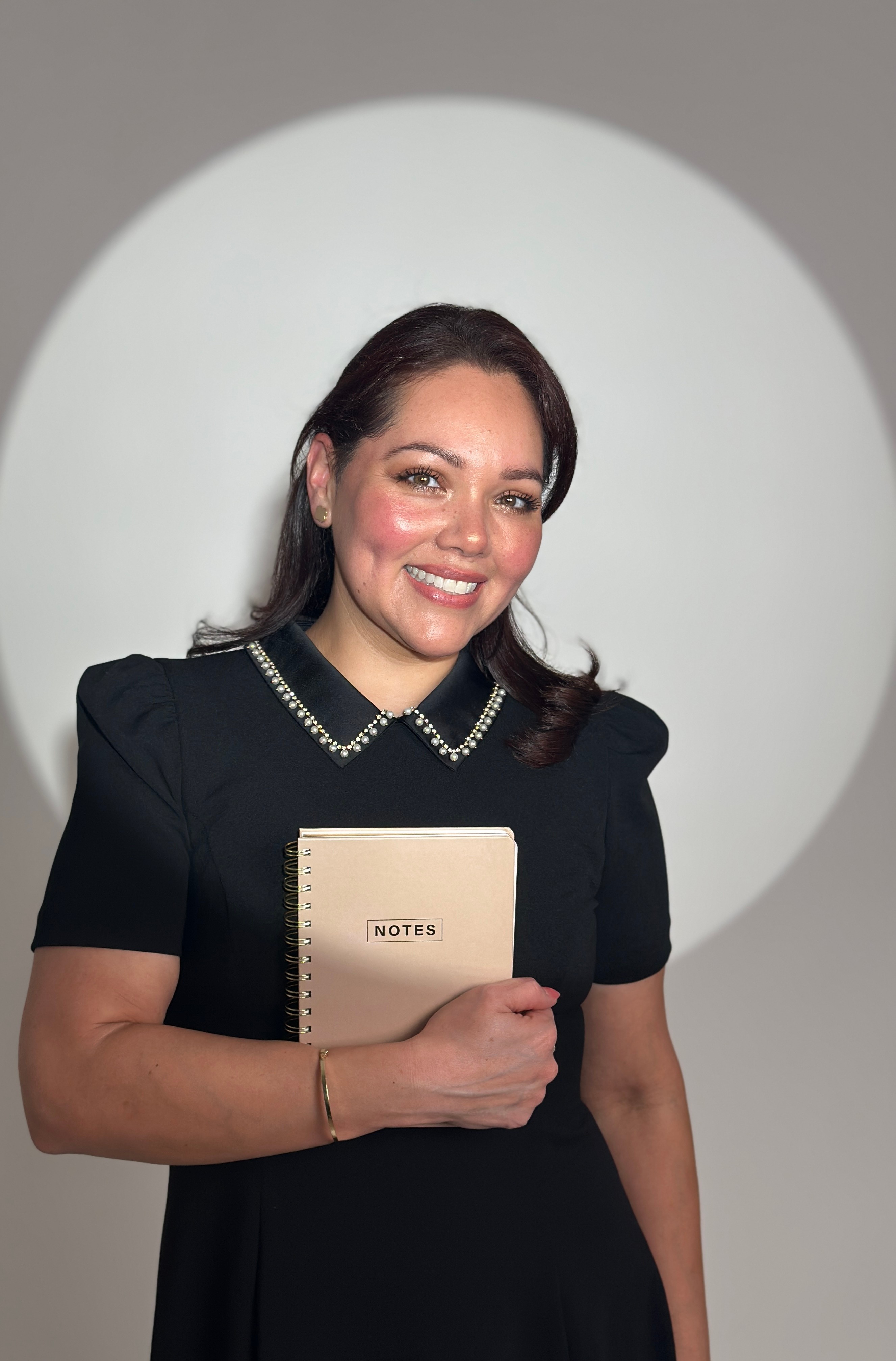 Smiling woman holding a notebook, standing in front of a circular light projection, wearing a black dress with pearl details.
