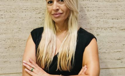 Woman with long blonde hair wearing a black sleeveless top, standing against a textured beige wall.