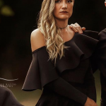 Woman with long wavy blonde hair wearing a black off-shoulder dress and choker necklace, standing indoors.