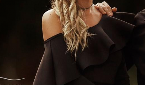 Woman with long wavy blonde hair wearing a black off-shoulder dress and choker necklace, standing indoors.
