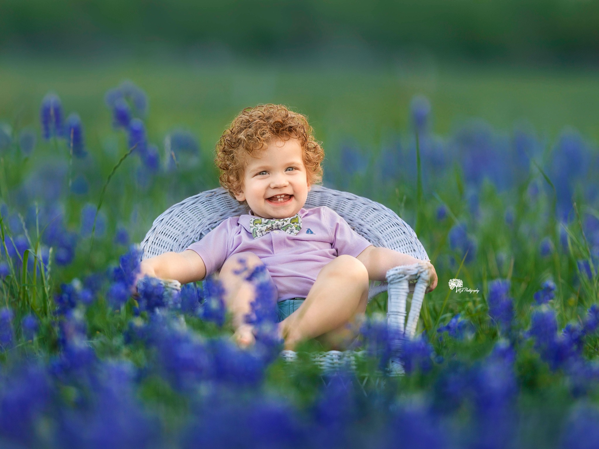 Child with curly hair smiling, sitting in a chair among blue flowers outdoors.