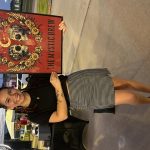 Woman standing next to a colorful poster with skulls and flowers, smiling, wearing a black top and checkered skirt, outdoors.