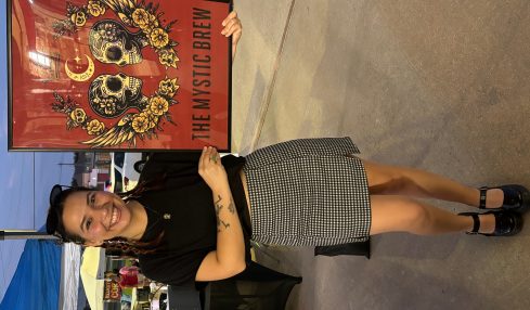 Woman standing next to a colorful poster with skulls and flowers, smiling, wearing a black top and checkered skirt, outdoors.