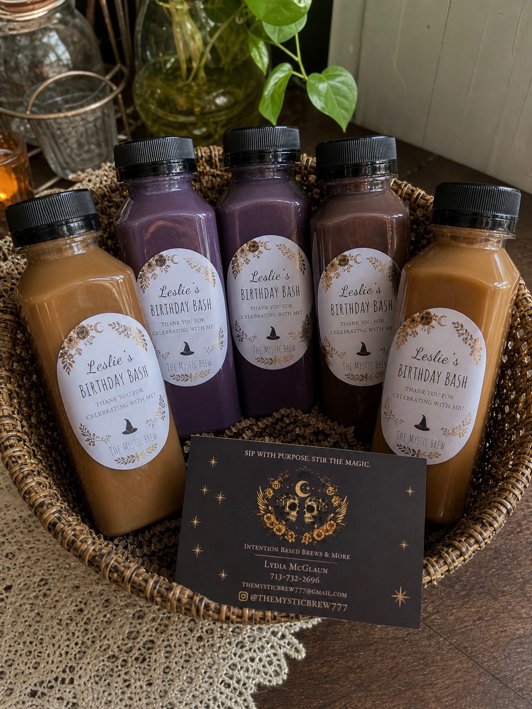 Five bottles of beverage in a basket, with a business card in front, plants and jars in background.