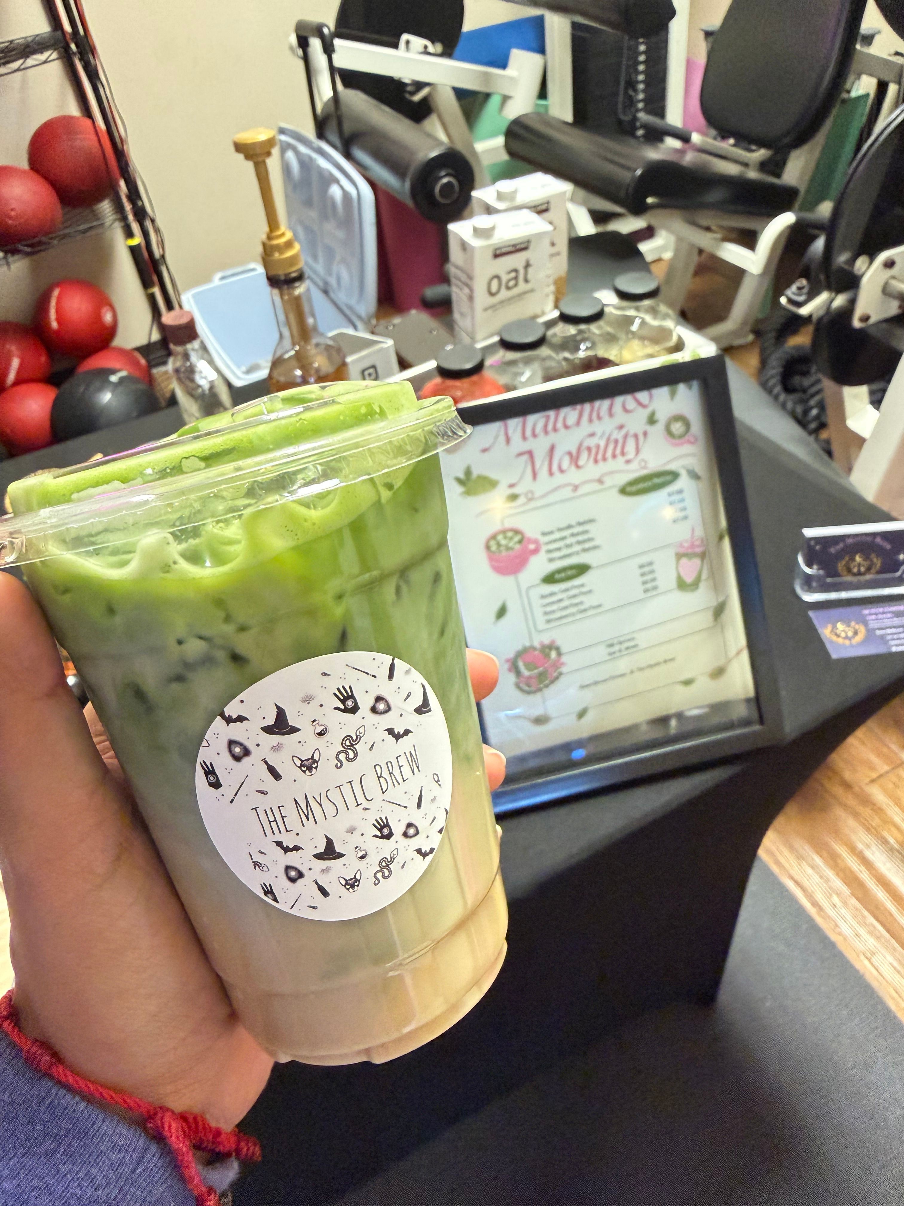 Hand holding a large iced matcha drink with a white circular sticker, in front of a tablet and gym equipment.