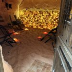 Indoor space with chairs, illuminated salt wall, sandy floor, and a partially open door on the right.