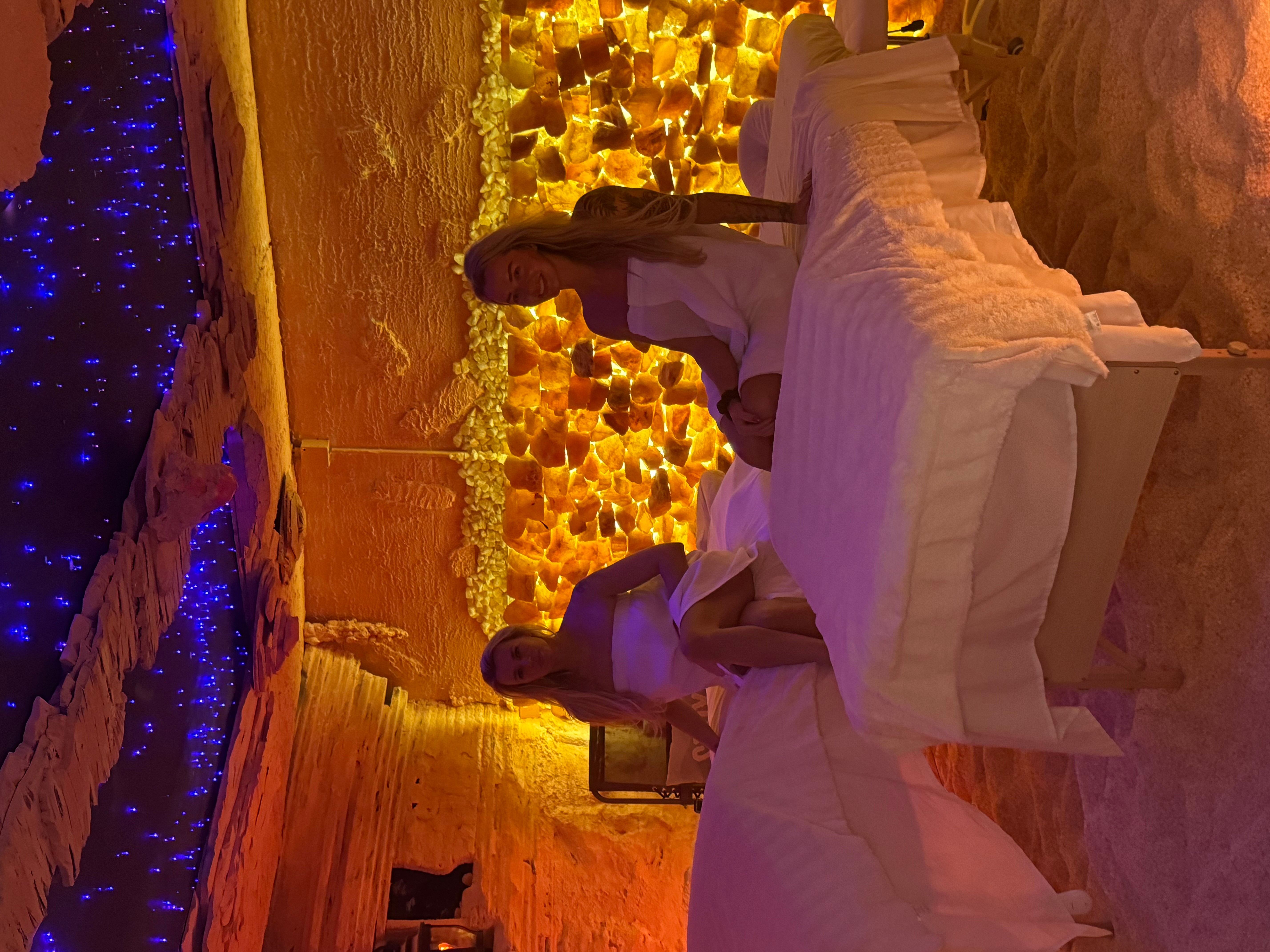 Two people sit at a table in a warmly lit, decorated indoor space with golden and blue lighting, and a textured wall.