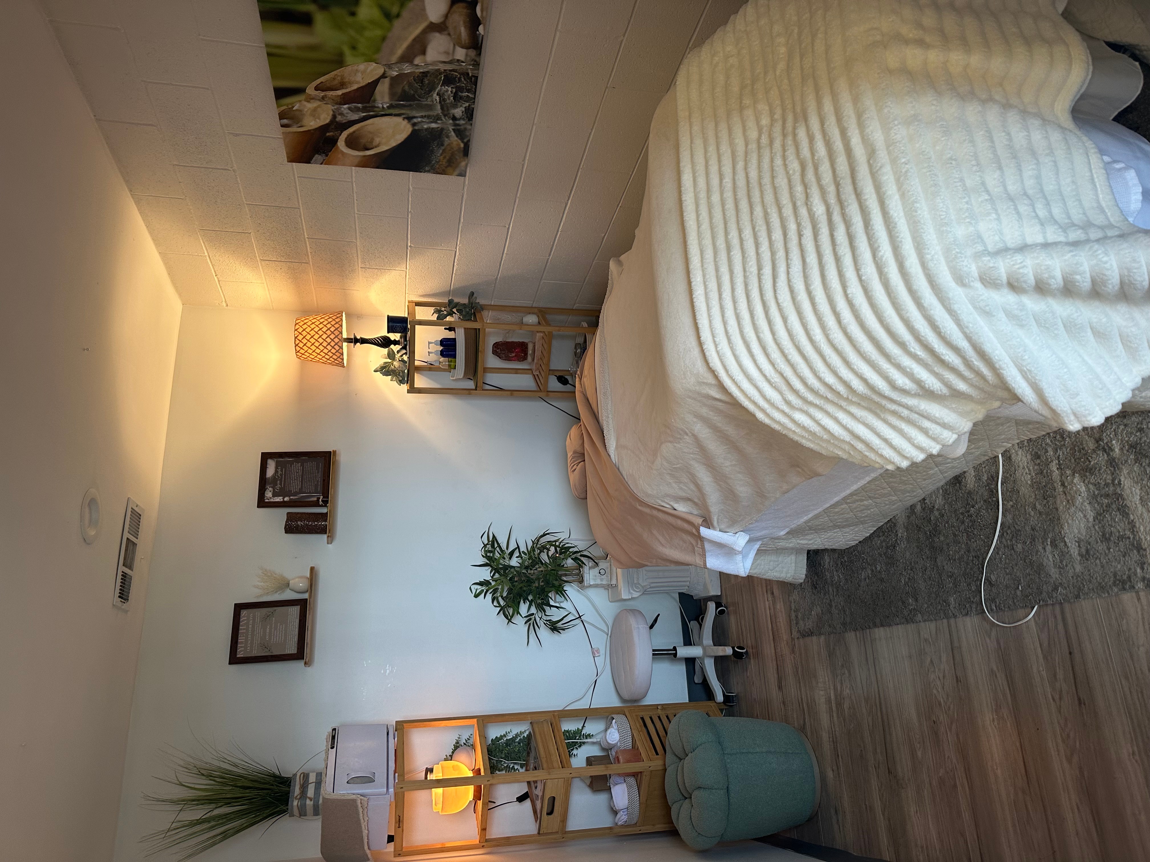 Bedroom with bed, nightstand, plant, and wall decorations, illuminated by a lamp, with wooden flooring.