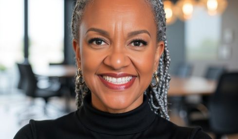 Smiling woman with gray hair styled in braids, wearing a black turtleneck, in a modern office setting with large windows and hanging lights.