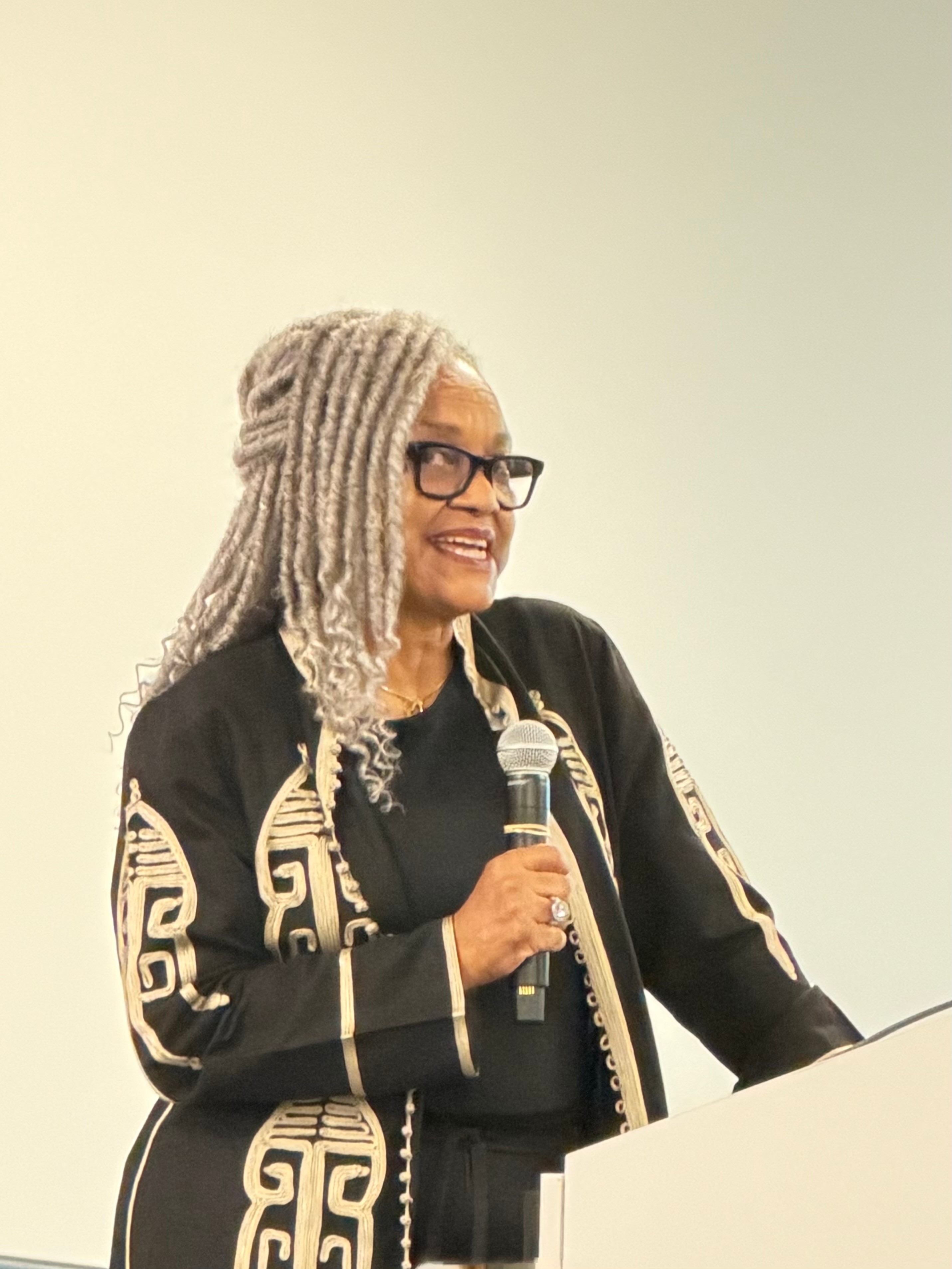 Woman with gray dreadlocks and glasses speaking into a microphone, standing behind a podium.