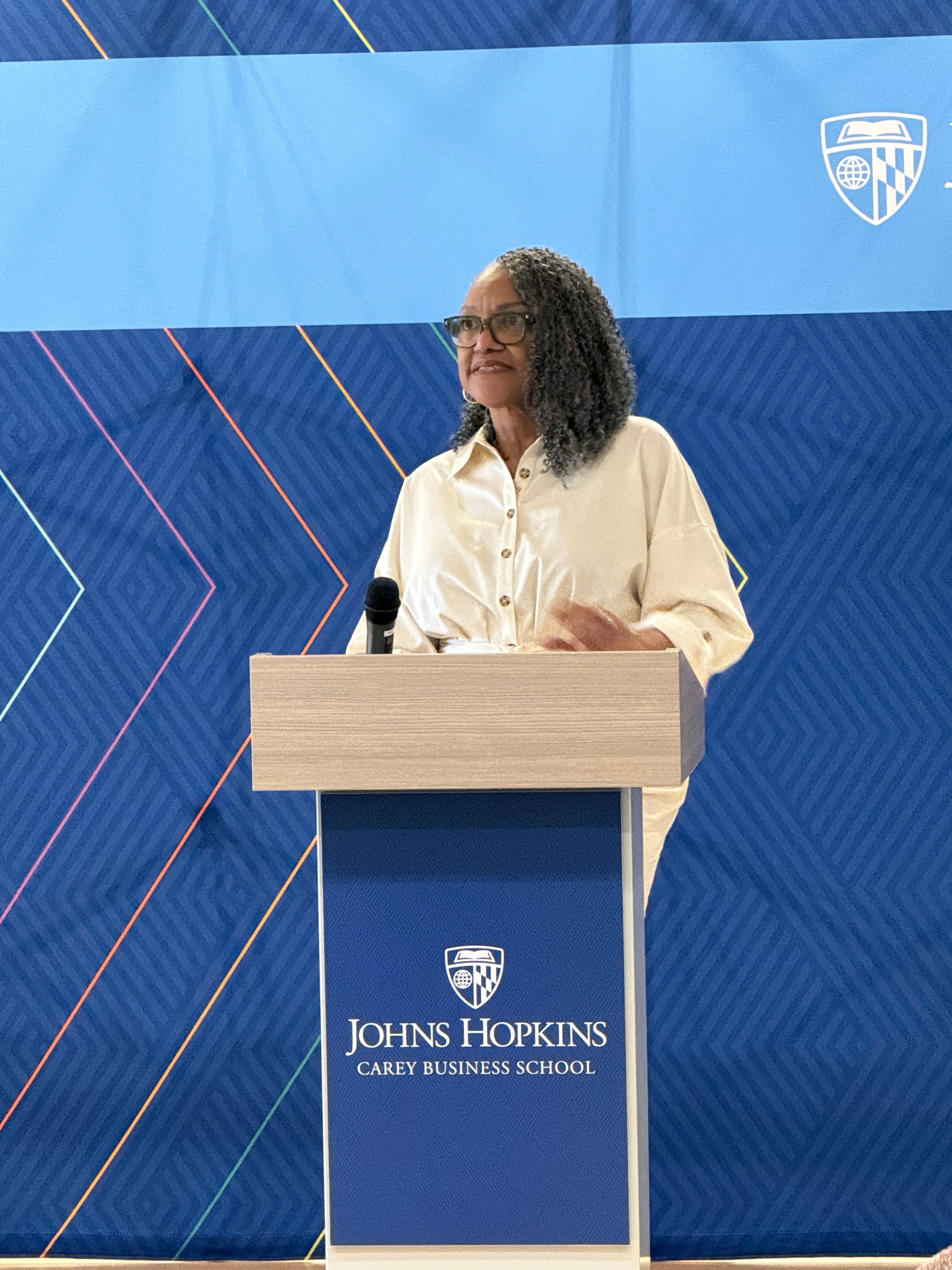 Woman speaking at a podium with a blue background and Johns Hopkins logo, facing left.