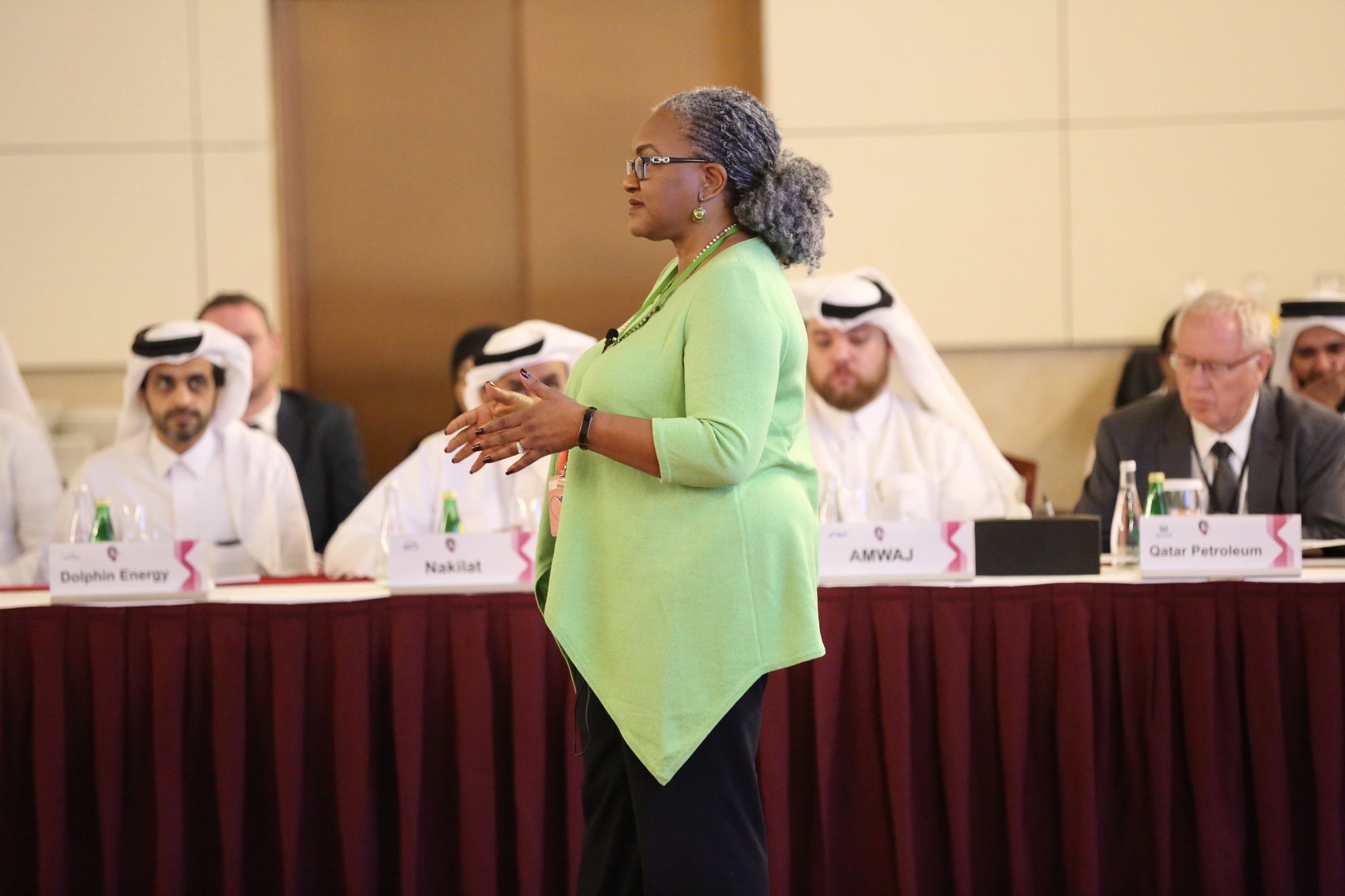 Woman standing and speaking at a conference table with seated men in traditional Middle Eastern attire and suits, in a conference room.