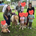 Group of children and two adults with a pony outdoors, some children holding plush toys, in a grassy area with trees.