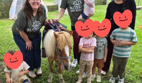 Group of children and two adults with a pony outdoors, some children holding plush toys, in a grassy area with trees.