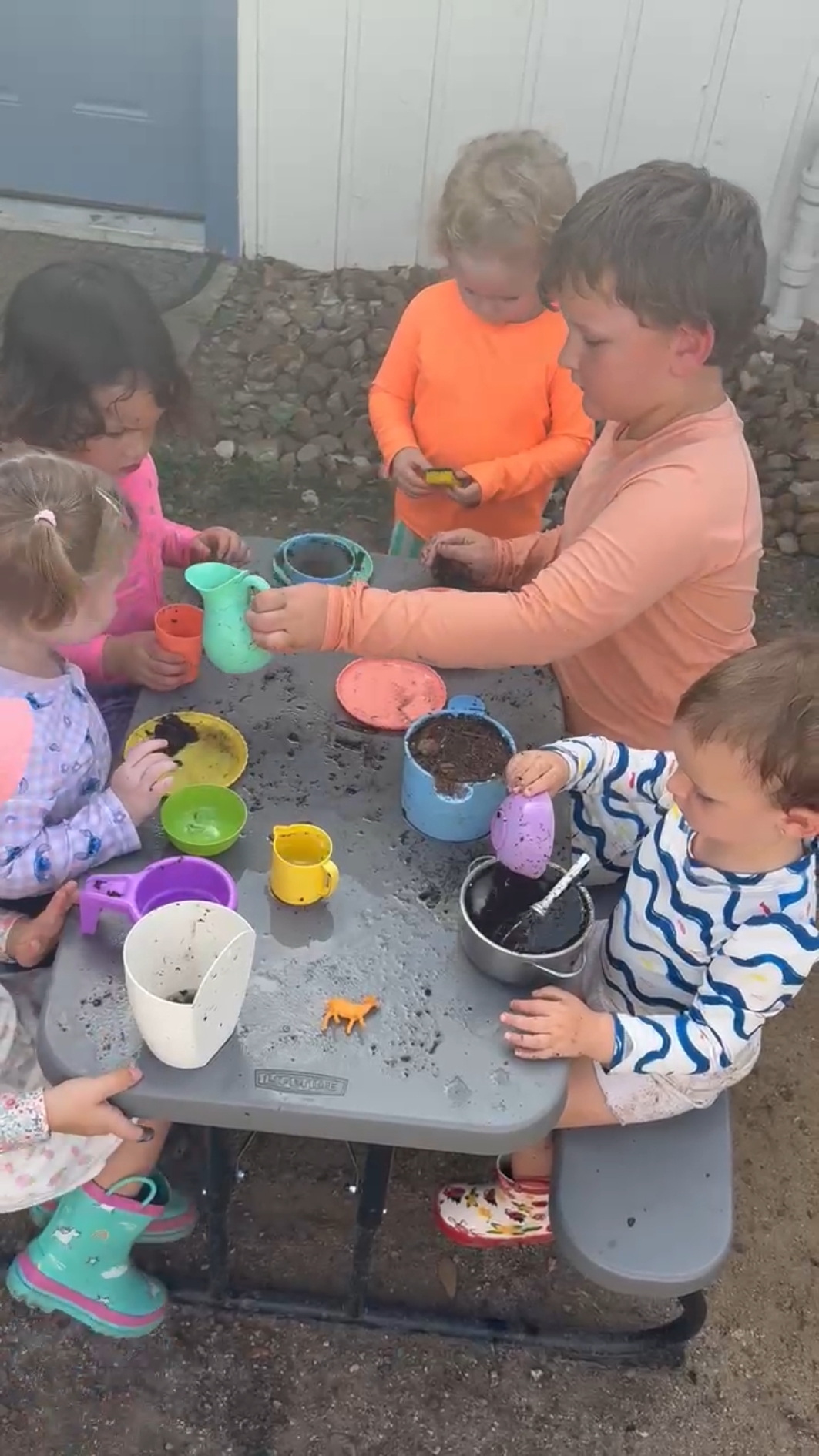 Children and an adult playing with colorful toys and containers at a table outdoors.