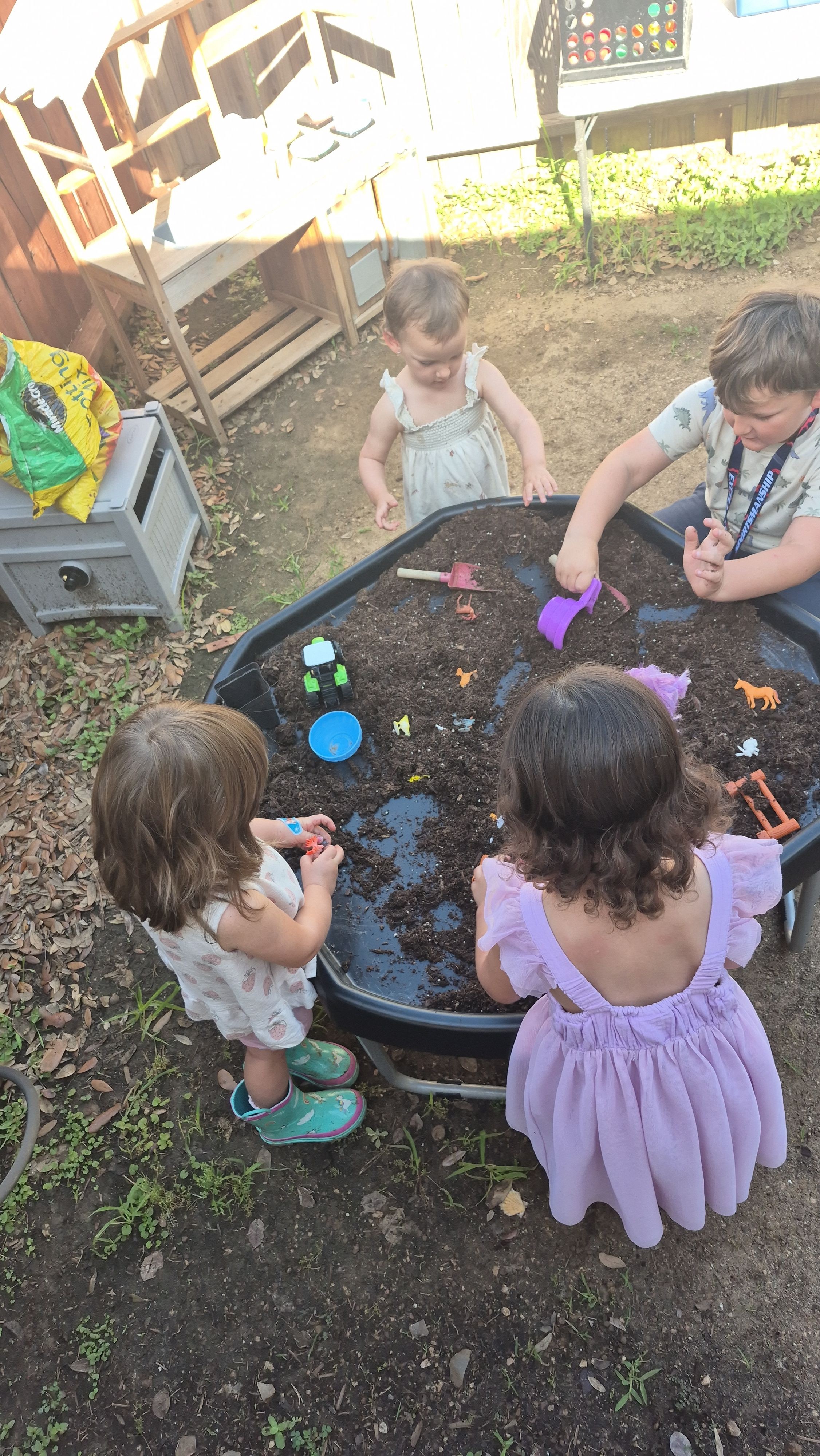 Children playing in a garden, some sitting and some standing, with toys and a small trampoline visible.