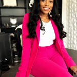 Woman in pink suit sitting on a desk, smiling, with a bookshelf and window in the background.