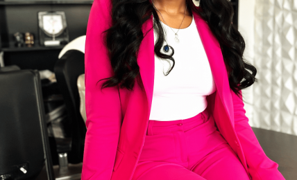 Woman in pink suit sitting on a desk, smiling, with a bookshelf and window in the background.