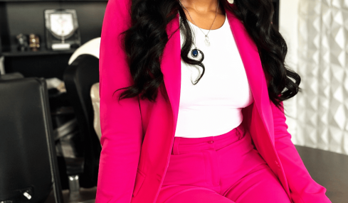 Woman in pink suit sitting on a desk, smiling, with a bookshelf and window in the background.
