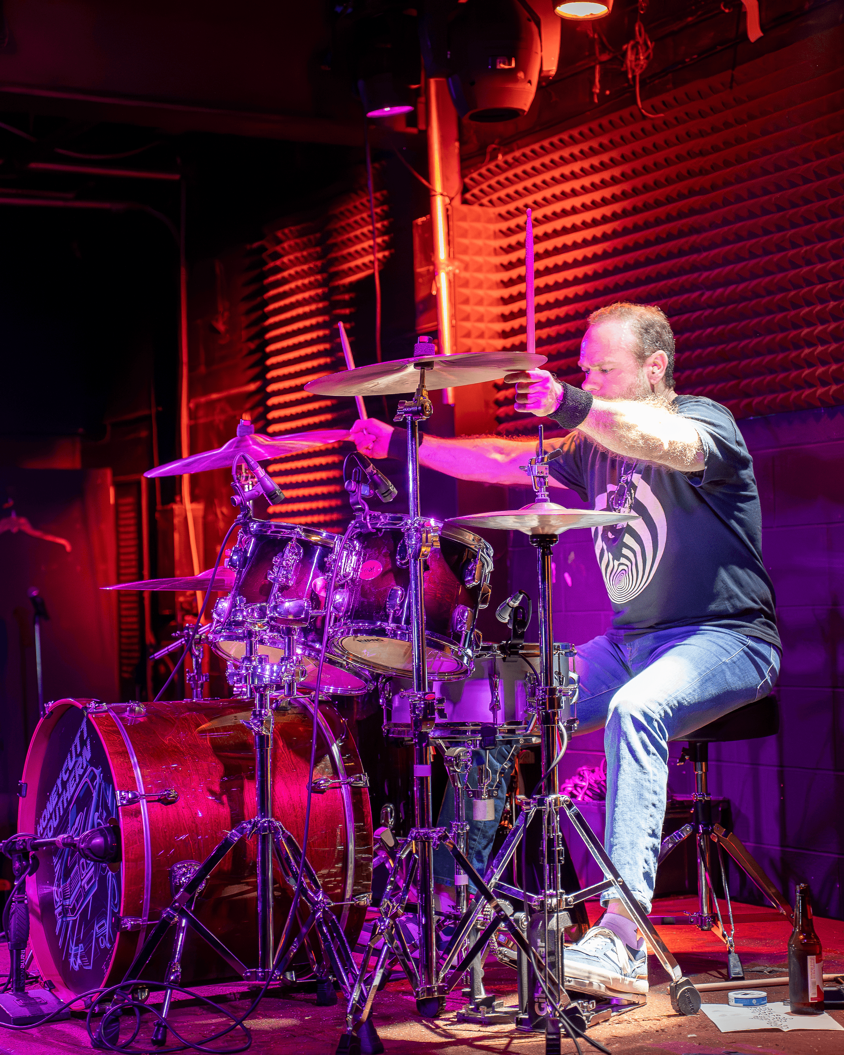 A man playing drums on stage with red and purple lighting, wearing a black vest and white pants.