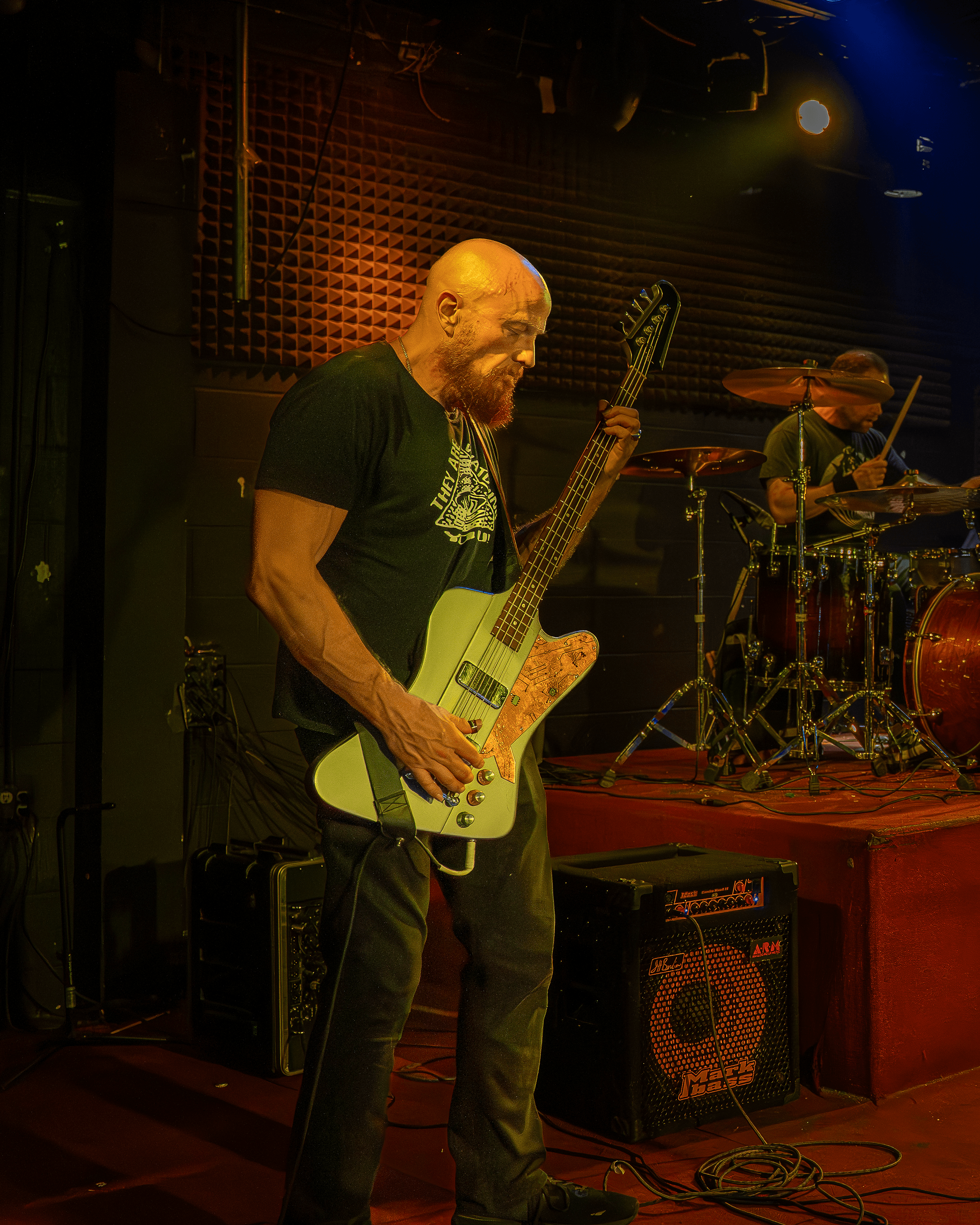 Bald man playing electric bass guitar on stage with drummer in background, stage lighting, and musical equipment.