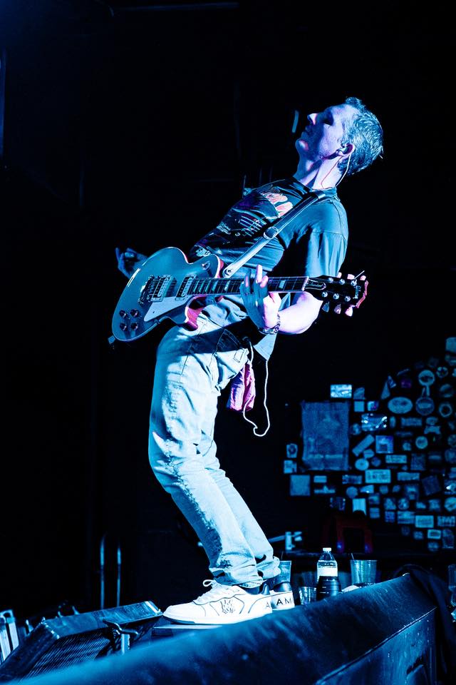 Person playing guitar on stage with dark background, illuminated by blue light, standing on a platform with drinks nearby.
