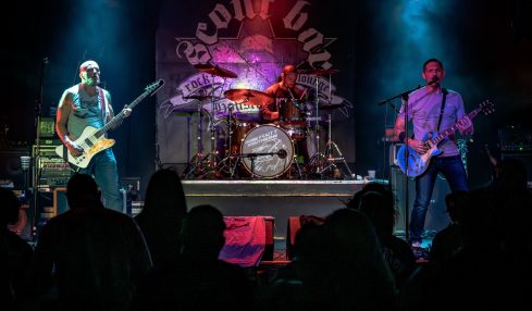 Band performs on stage with three musicians playing guitar and drums, audience silhouettes in foreground, stage backdrop with logo.