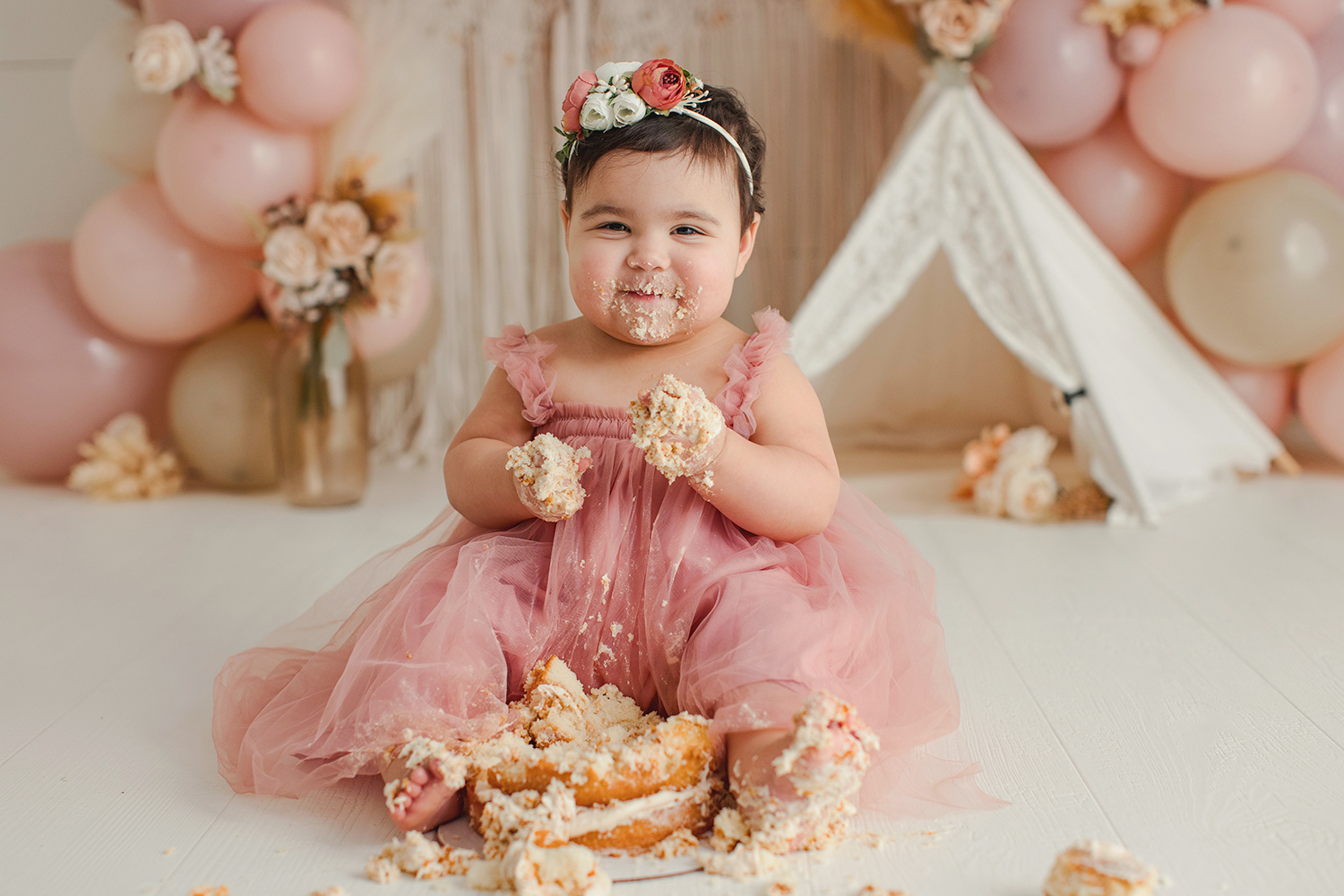 Smiling young girl in pink dress sitting on floor with balloons and a tent in background.