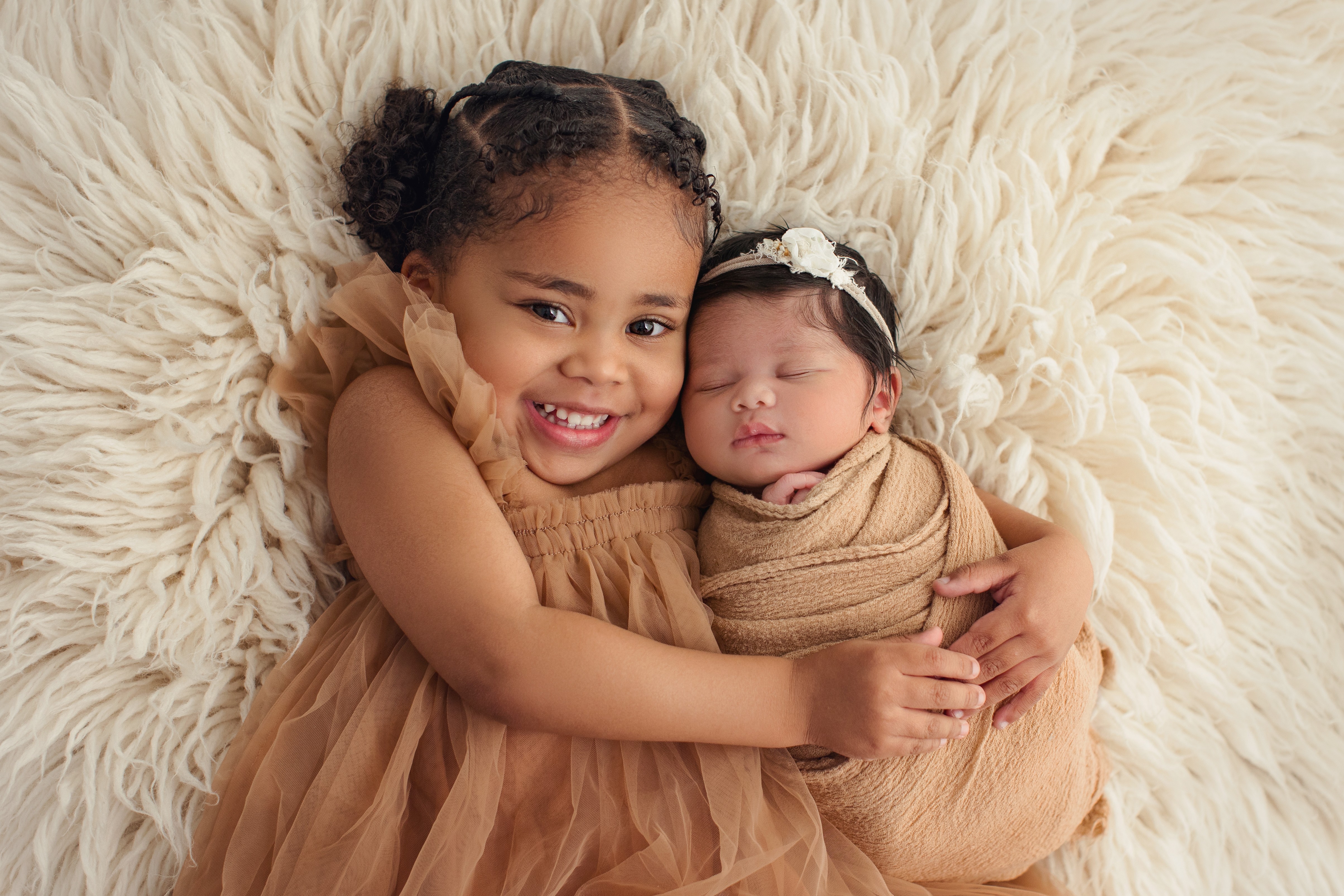 Two young children, one smiling girl and a sleeping baby, lying on a fluffy cream-colored surface, hugging each other.