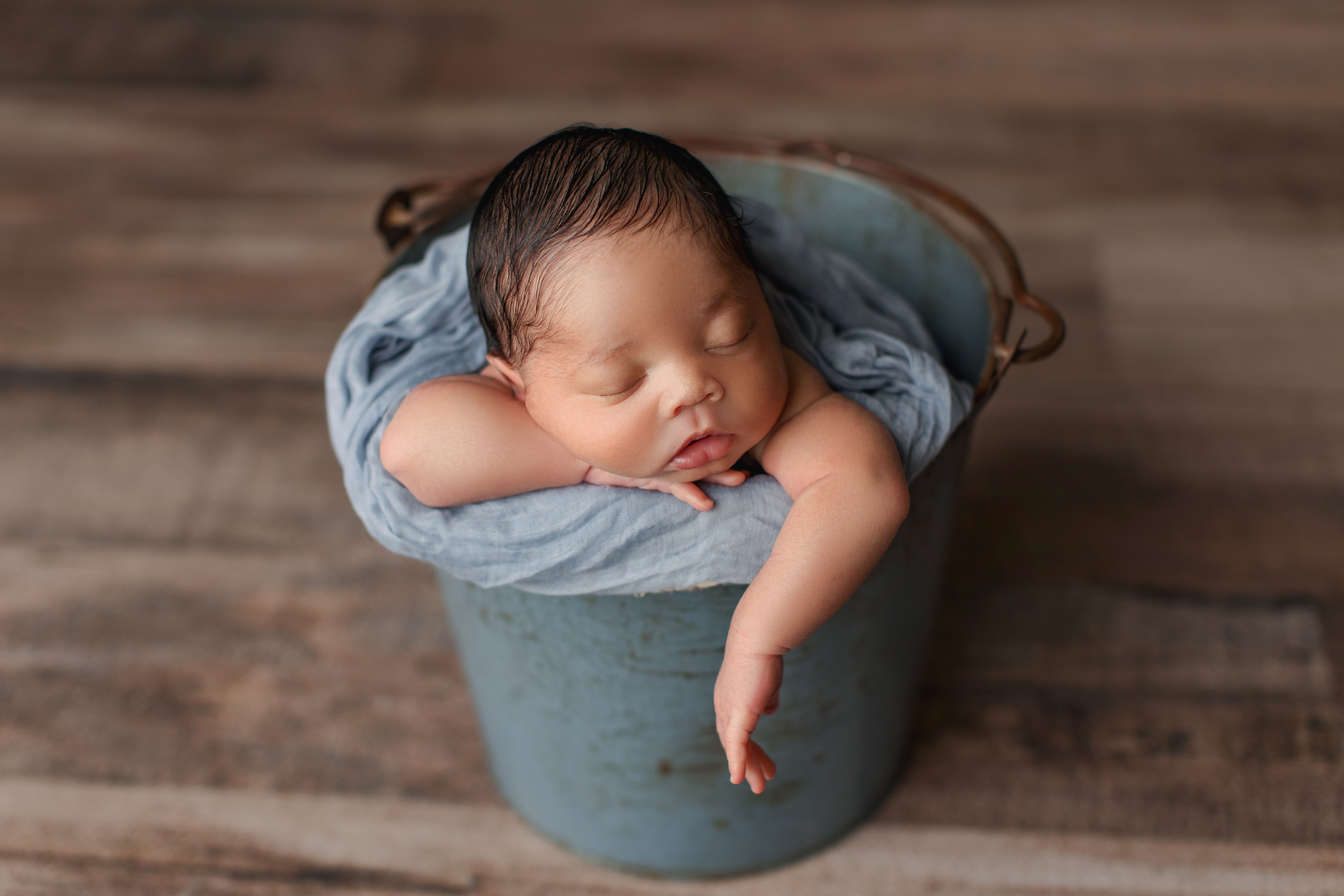 Sleeping baby in a small bucket, resting with one arm hanging over the side, on a wooden floor.