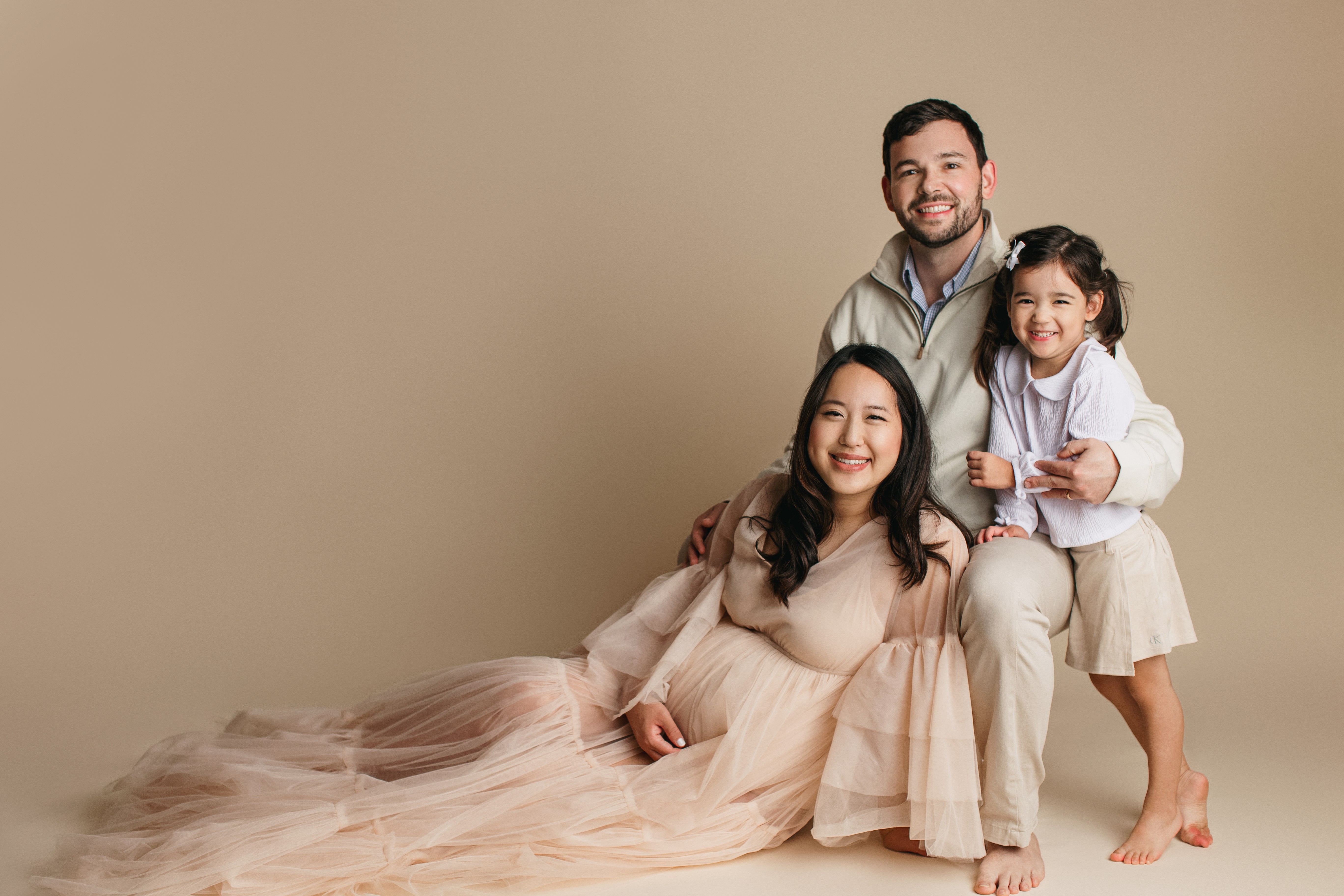 Family of three smiling, sitting and standing against a beige background, woman in a light dress, man in a shirt, girl in a white dress.