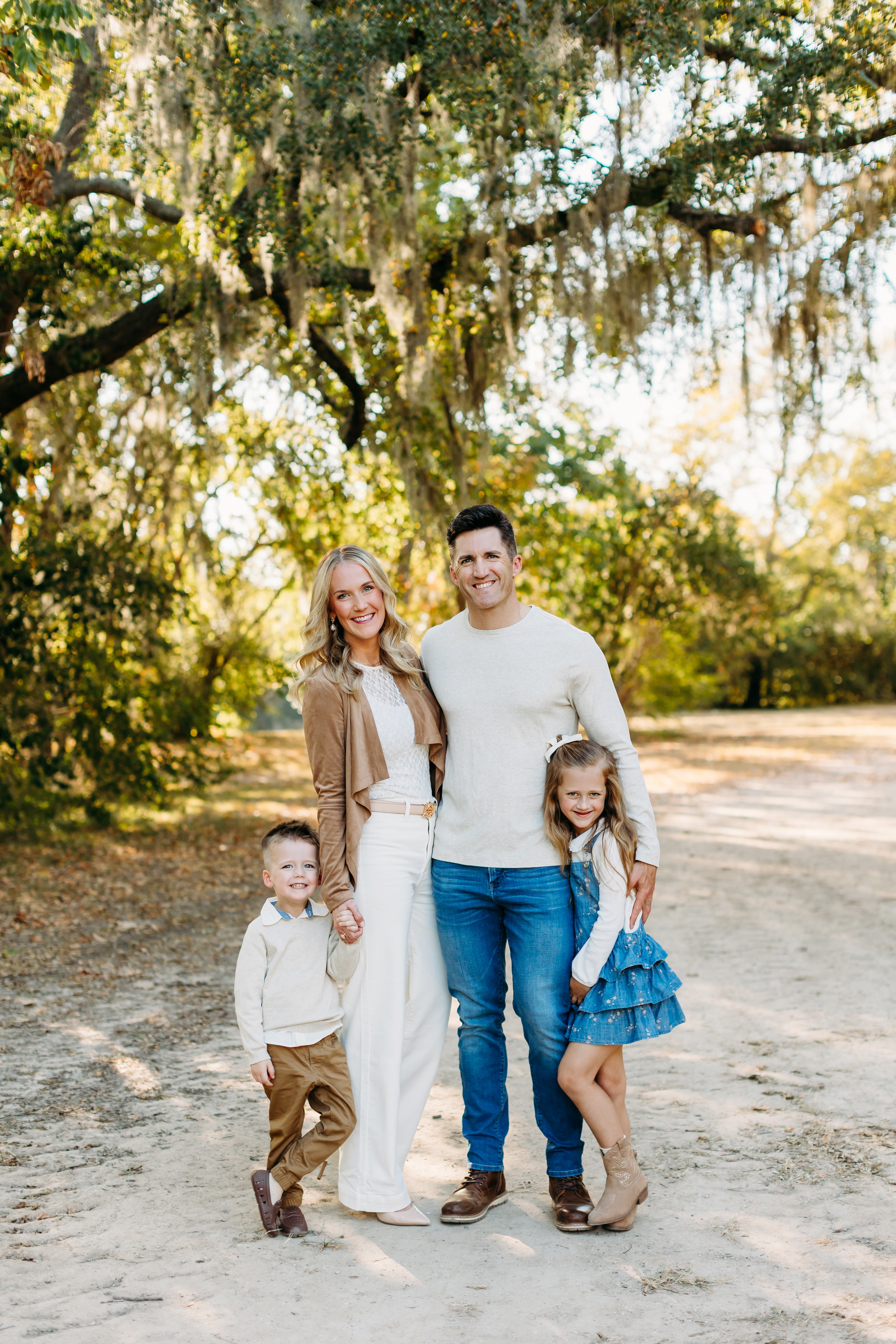 Family of four standing outdoors on a dirt path with trees and sunlight in background, smiling, with two children and two adults.